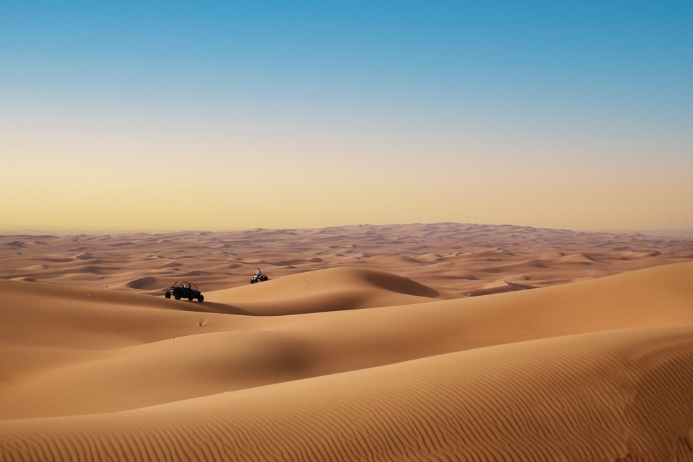 Vastidão do deserto de Dubai com dunas douradas ondulantes sob um céu azul gradiente; um buggy e uma moto são vistos ao longe.