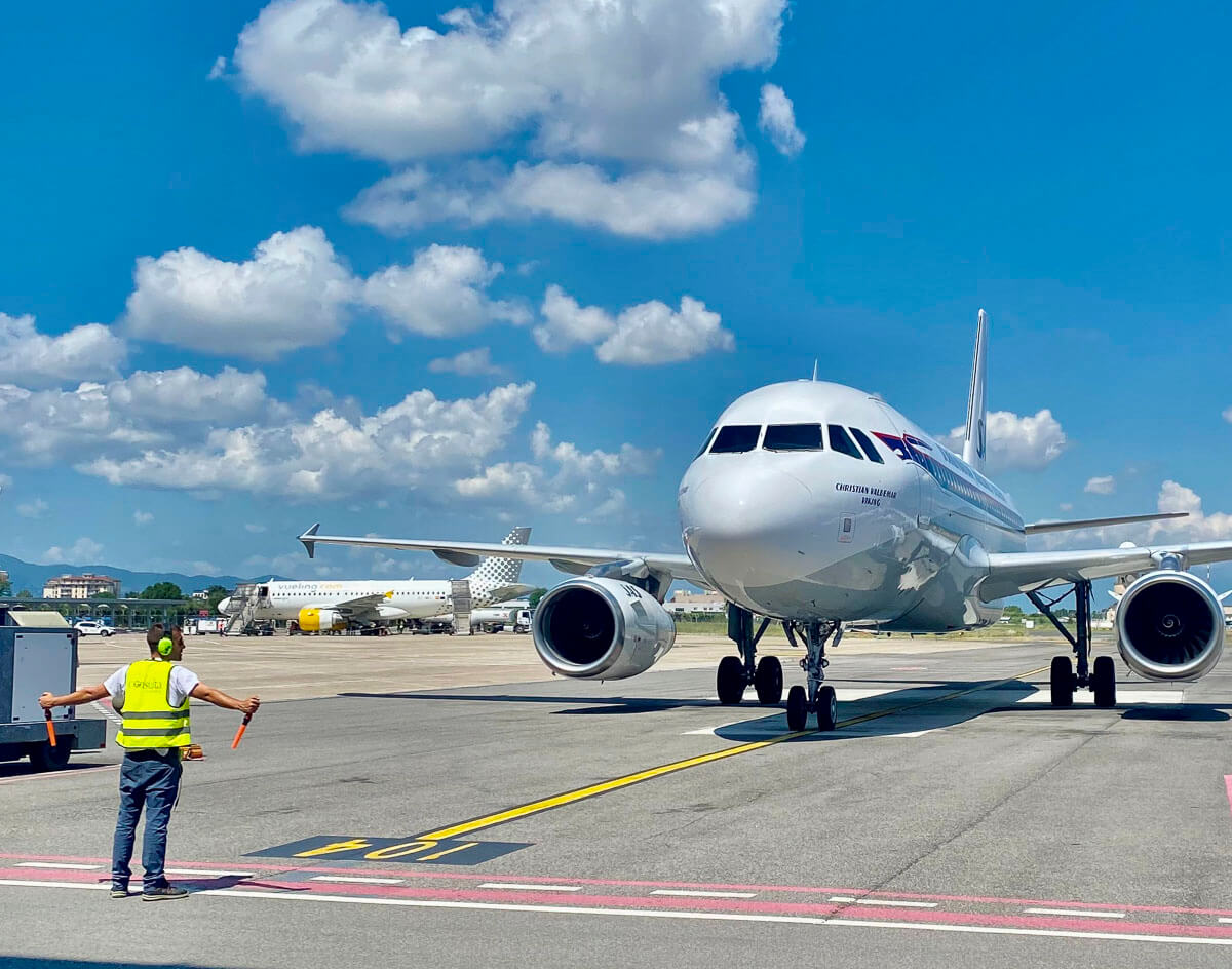 Avião sendo guiado para o terminal por funcionário com colete refletivo no Aeroporto Amerigo Vespucci, em um dia ensolarado com céu azul e algumas nuvens.