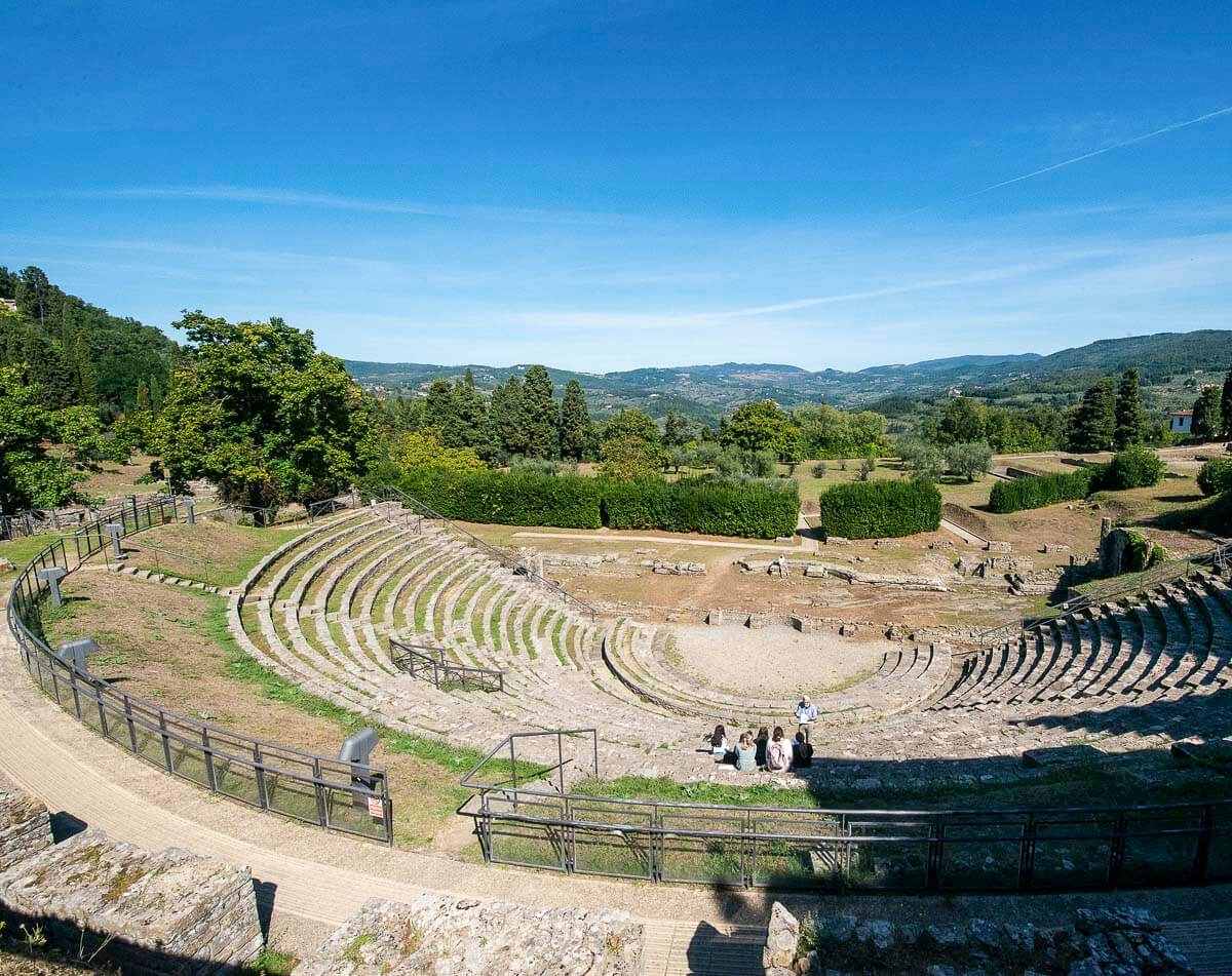 Ruínas de um antigo anfiteatro romano em Fiesole, nos arredores de Florença, cercado por vegetação e colinas.