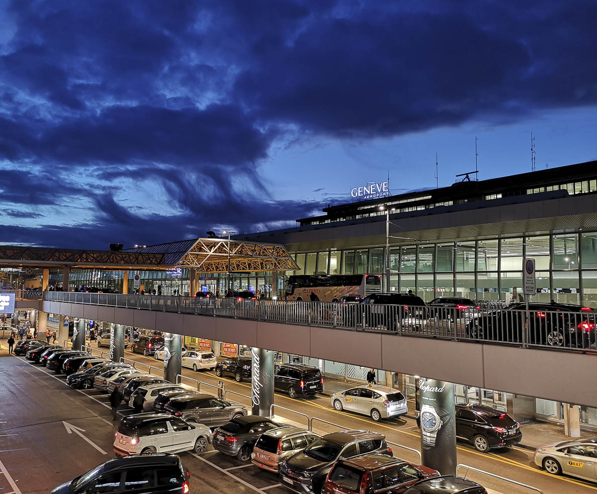 Vista externa do Aeroporto Internacional de Genebra à noite, com carros estacionados e um céu escuro ao fundo.