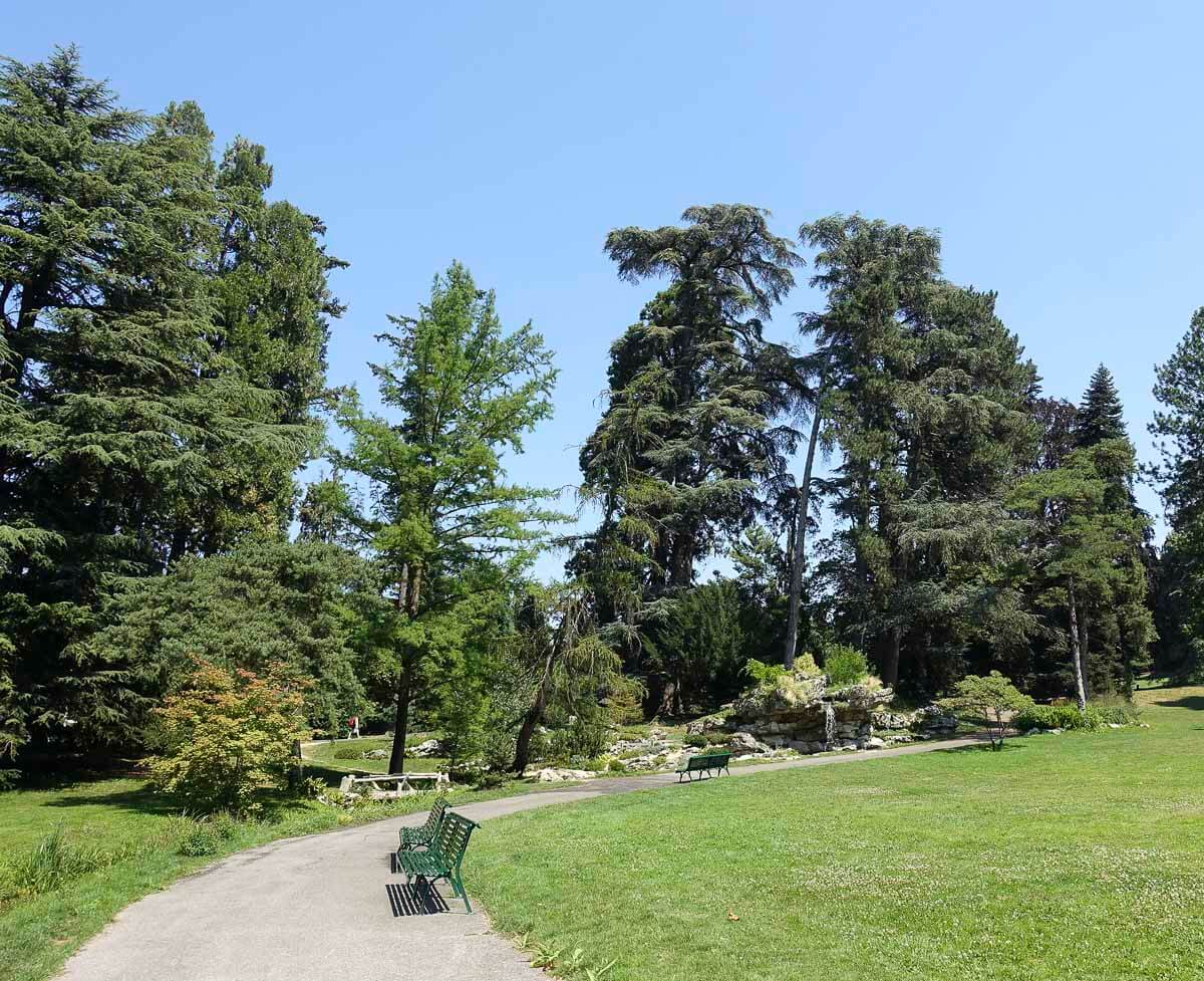 Caminho com bancos e vegetação exuberante no Parc des Eaux-Vives, em Genebra, com céu azul ao fundo.