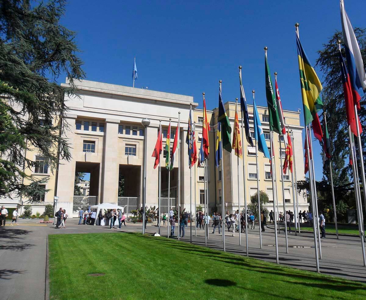 Fachada do Palais des Nations, sede da ONU em Genebra, com várias bandeiras de diferentes países ao longo do caminho.