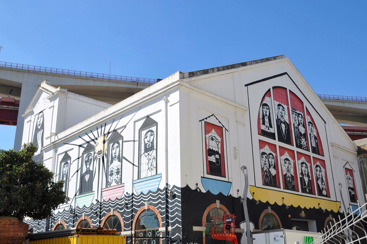Fachada do prédio do LX Factory, em Lisboa, com um mural colorido que retrata figuras estilizadas em janelas e barcos.
