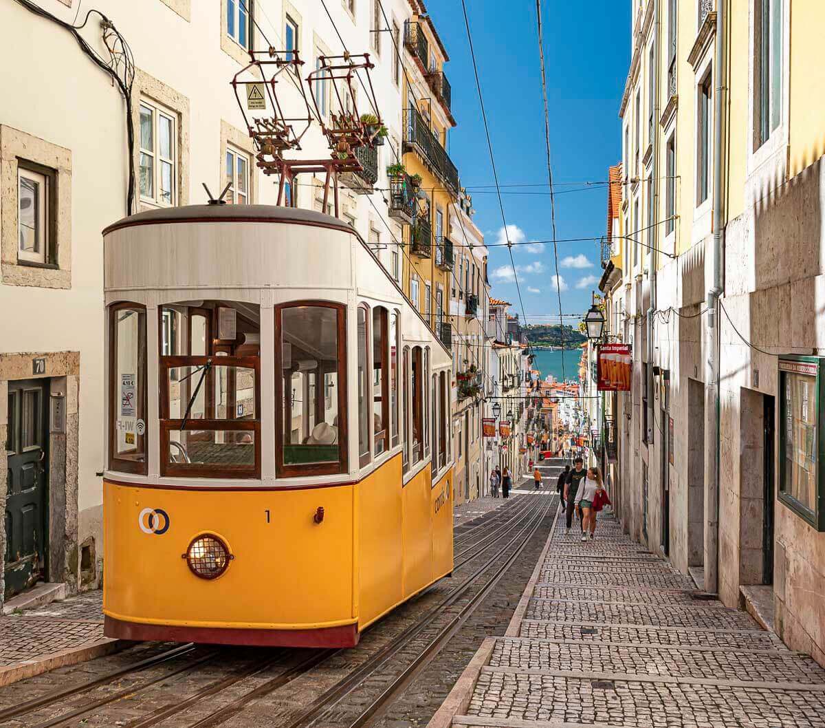 Bonde amarelo subindo uma rua íngreme no bairro da Bica, em Lisboa, com vista para o rio Tejo ao fundo.