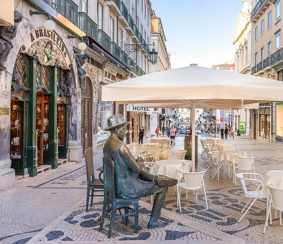 A fachada do café A Brasileira no bairro Chiado, com mesas externas e uma estátua de Fernando Pessoa sentado em uma cadeira.