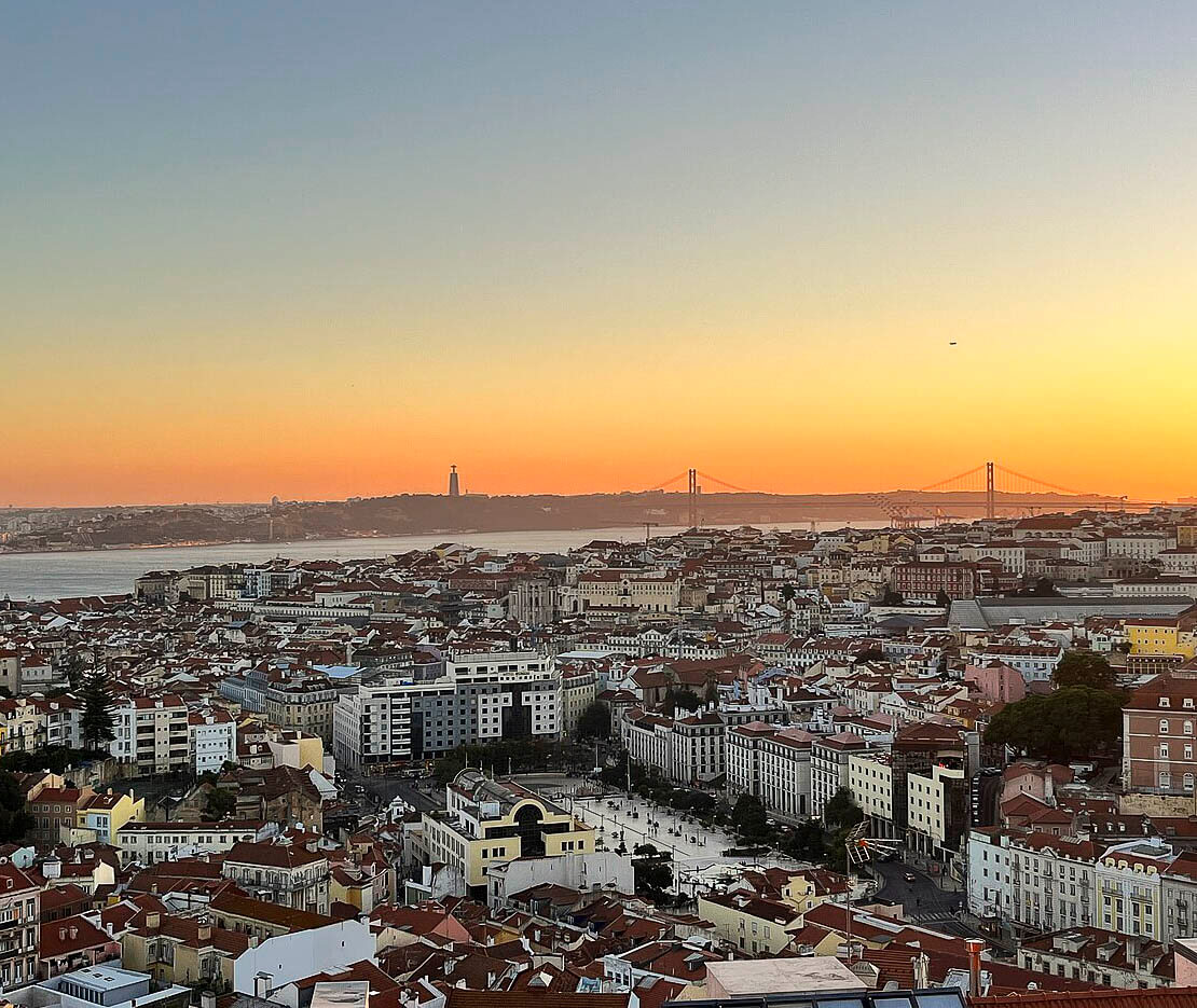 Vista panorâmica de Lisboa ao pôr do sol, com o rio Tejo ao fundo e a Ponte 25 de Abril destacada no horizonte.