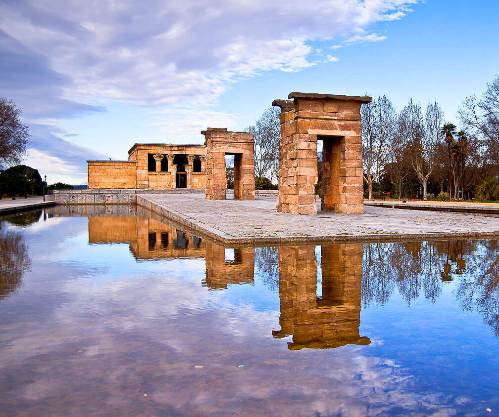 Templo de Debod, um antigo templo egípcio que o Egito deu de presente à Madrid, cercado por uma piscina refletindo suas estruturas de pedra sob um céu parcialmente nublado, com árvores ao fundo.