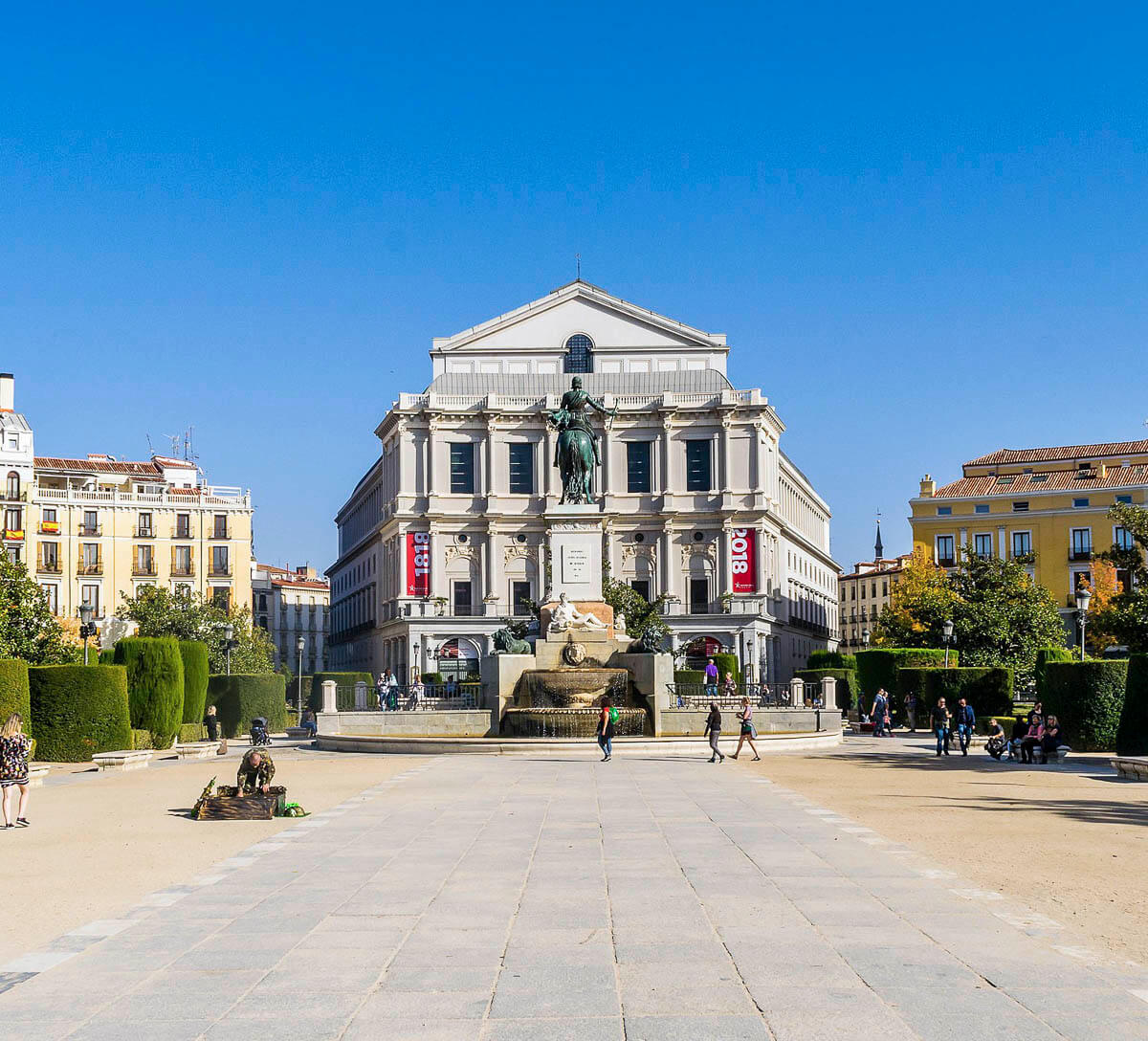 Praça em Madrid com o edifício do Teatro Real ao fundo, e turistas caminhando ao redor.