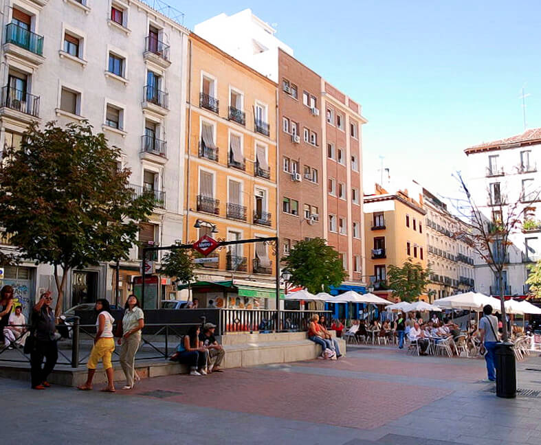 Praça em Madrid, com edifícios coloridos ao fundo, pessoas caminhando e cafés ao ar livre.
