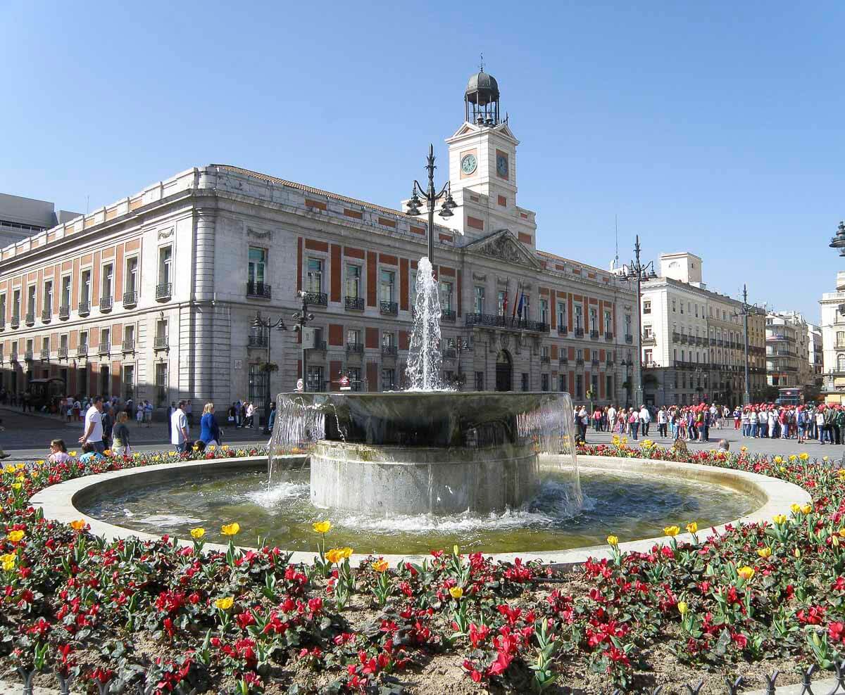 Fonte cercada por flores vermelhas e amarelas na praça Puerta del Sol, em Madrid, com o edifício histórico da Real Casa de Correos ao fundo e pessoas passeando pela área sob um céu claro.