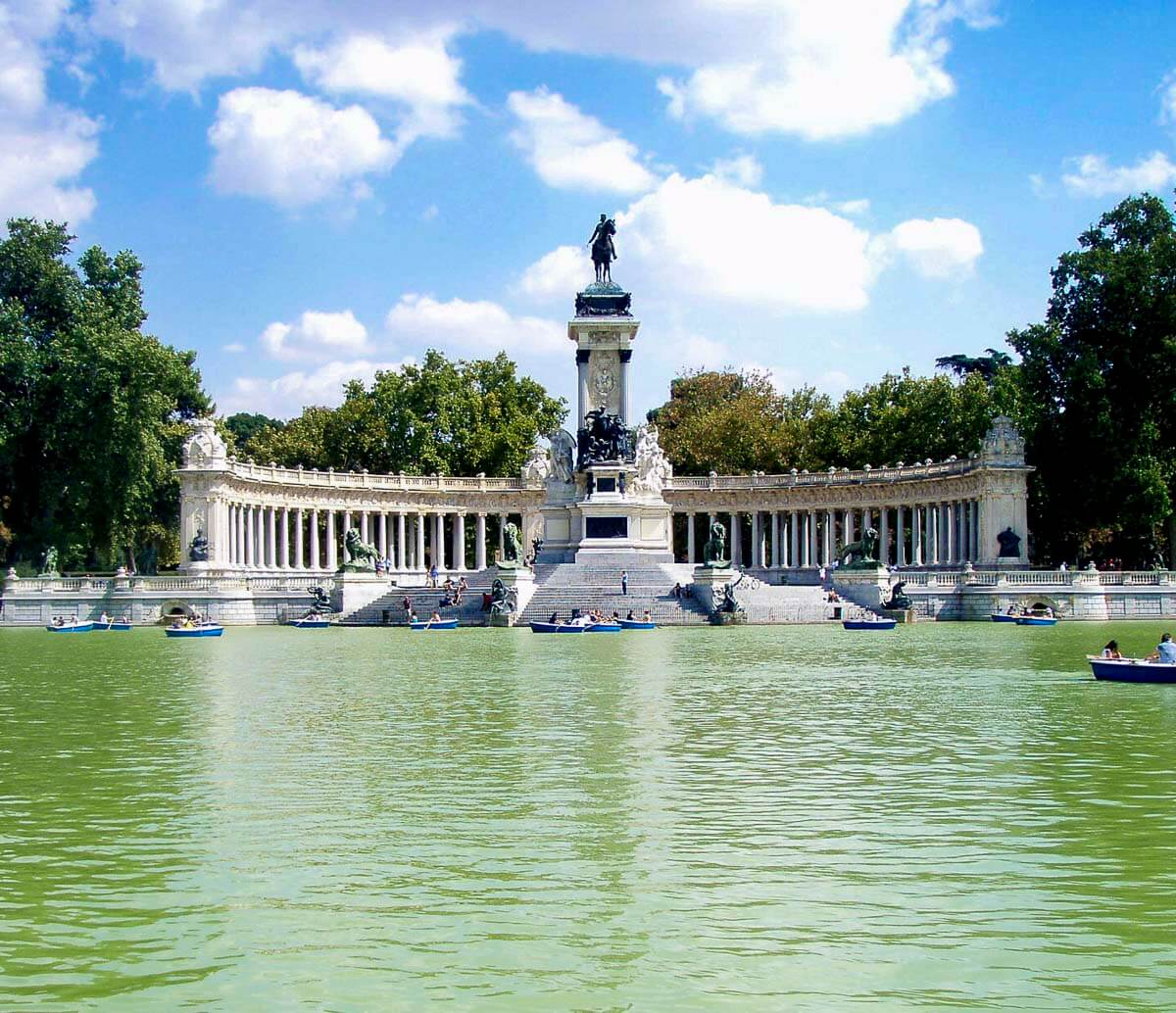 Monumento a Alfonso XII no Parque do Retiro, Madrid, com colunas brancas refletidas no grande lago em frente, sob um céu azul claro.