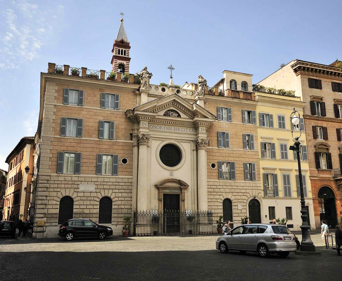 Fachada da histórica Igreja de Santa Brígida na Piazza Farnese, uma praça em Campo de' Fiori, com alguns carros estacionados e poucas pessoas caminhando.