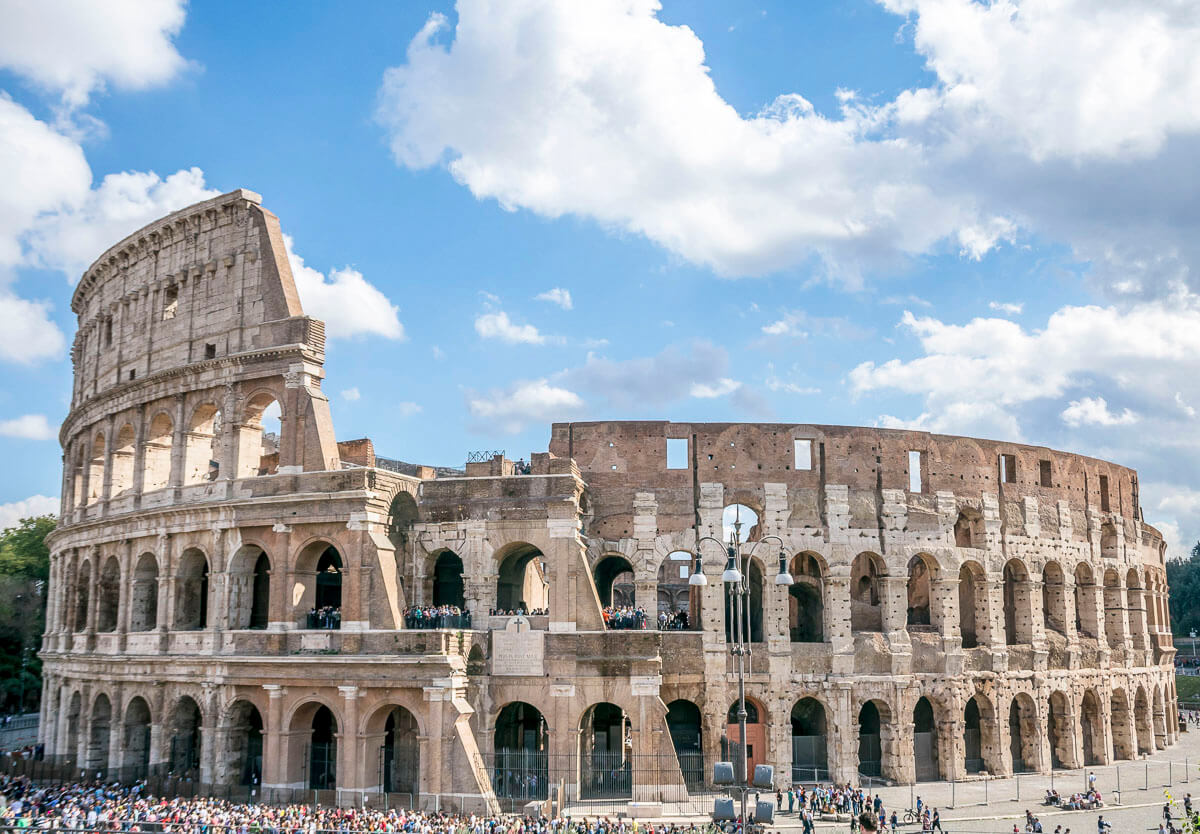 O Coliseu de Roma sob um céu azul com nuvens, com turistas ao redor e dentro do monumento.