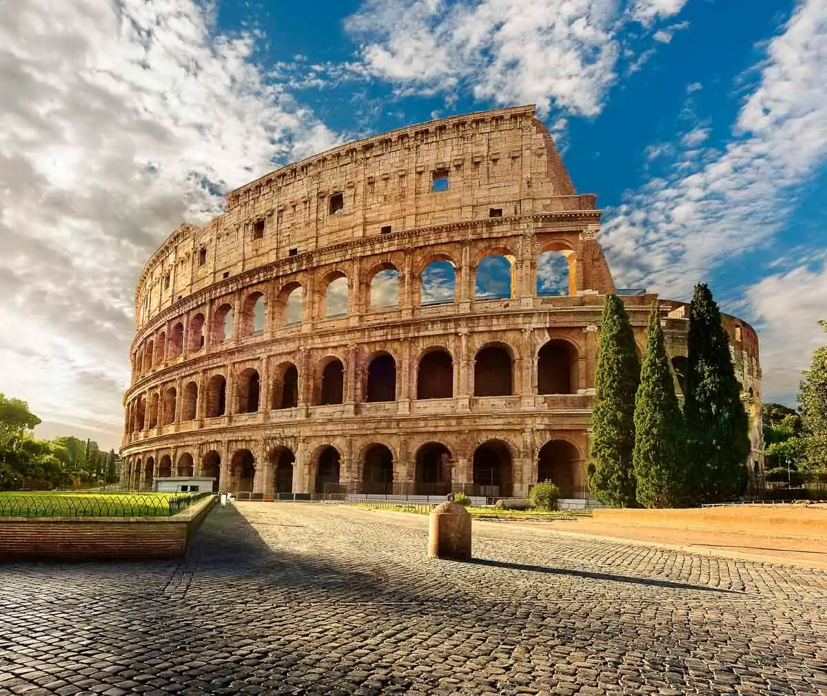 O Coliseu de Roma, sob um céu azul com nuvens, cercado por árvores e com calçamento de pedras ao redor.