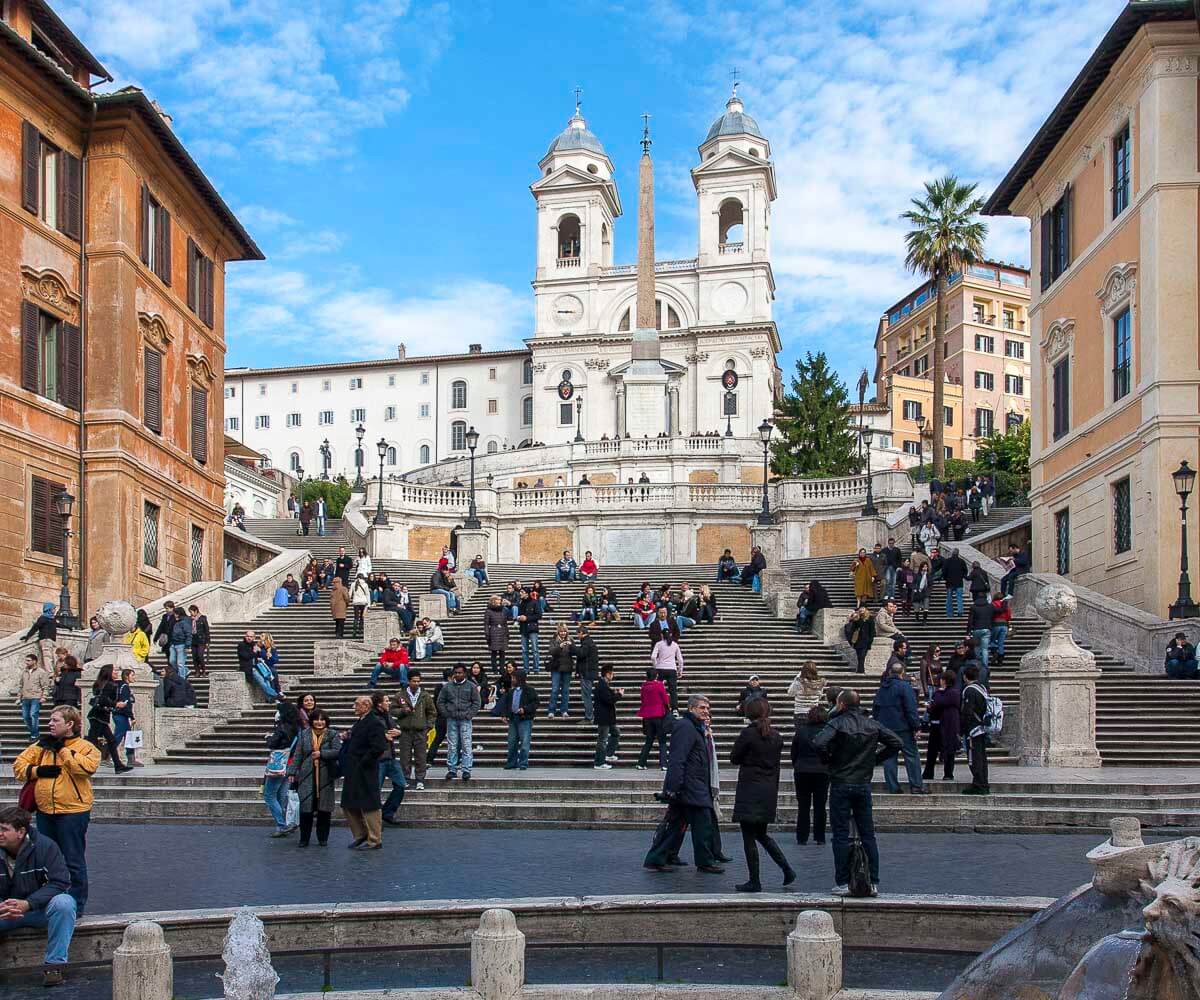 Escadaria da Piazza di Spagna em Roma com muitas pessoas sentadas e passeando, tendo ao fundo a Igreja Trinità dei Monti.