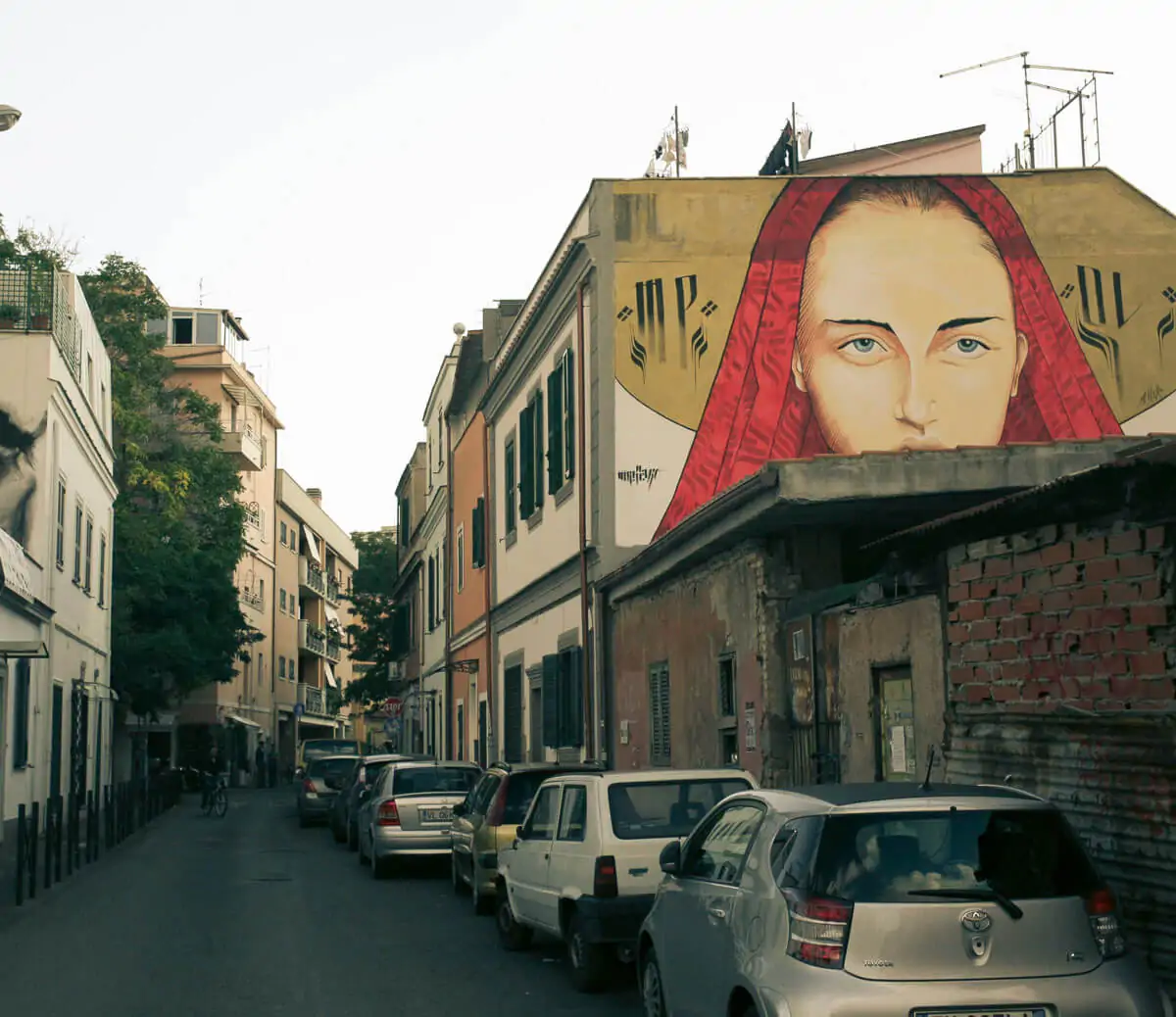 Rua estreita no bairro de Pigneto, Roma, com um grande mural de arte urbana retratando o rosto de uma mulher.