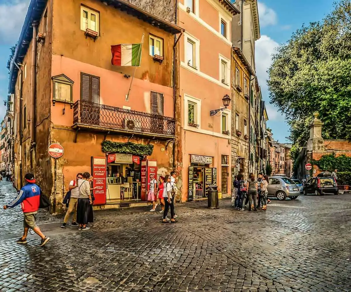 Esquina de uma rua em Trastevere, Roma, com fachadas de lojinhas em um prédio e bandeira da Itália pendurada na fachada.