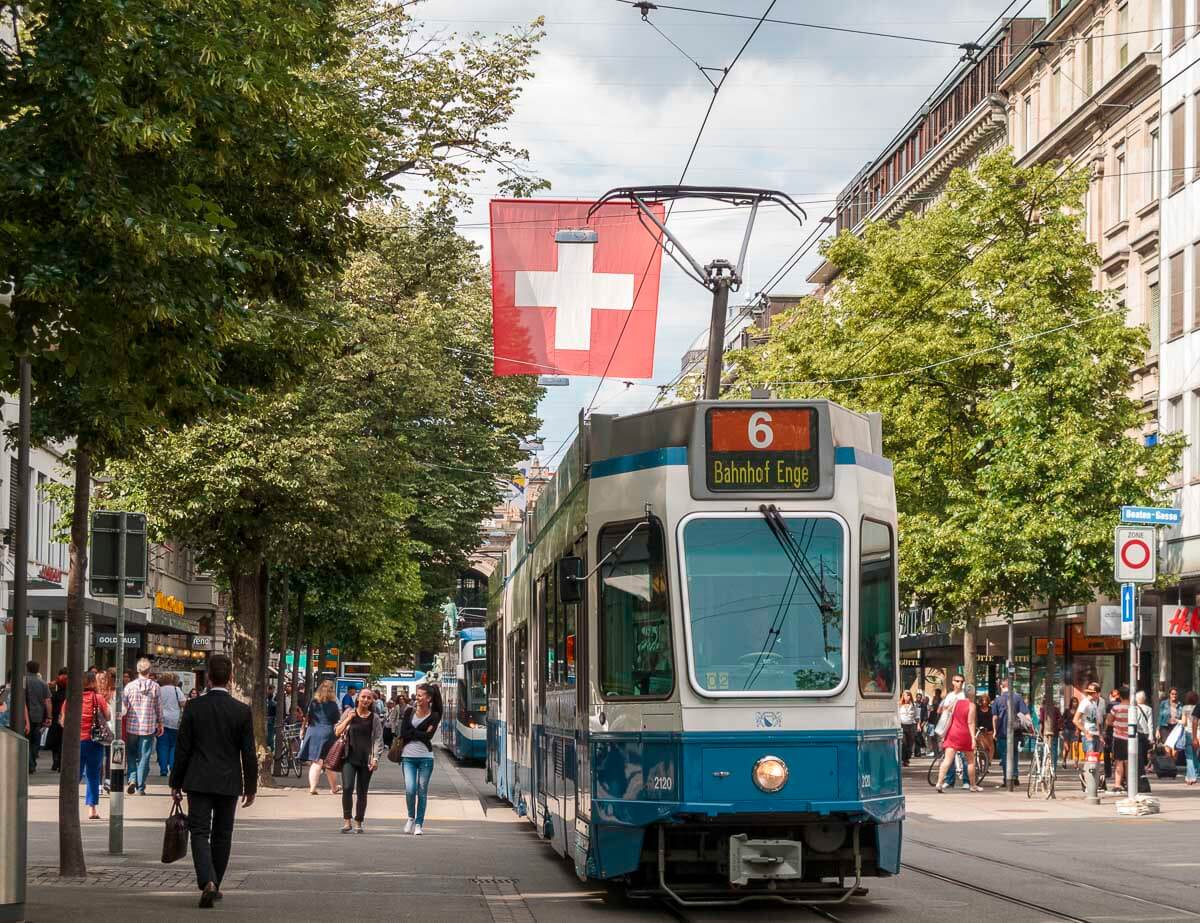 Bonde azul circulando pela Bahnhofstrasse, no centro de Zurique, com pedestres nas calçadas e uma bandeira da Suíça ao fundo.