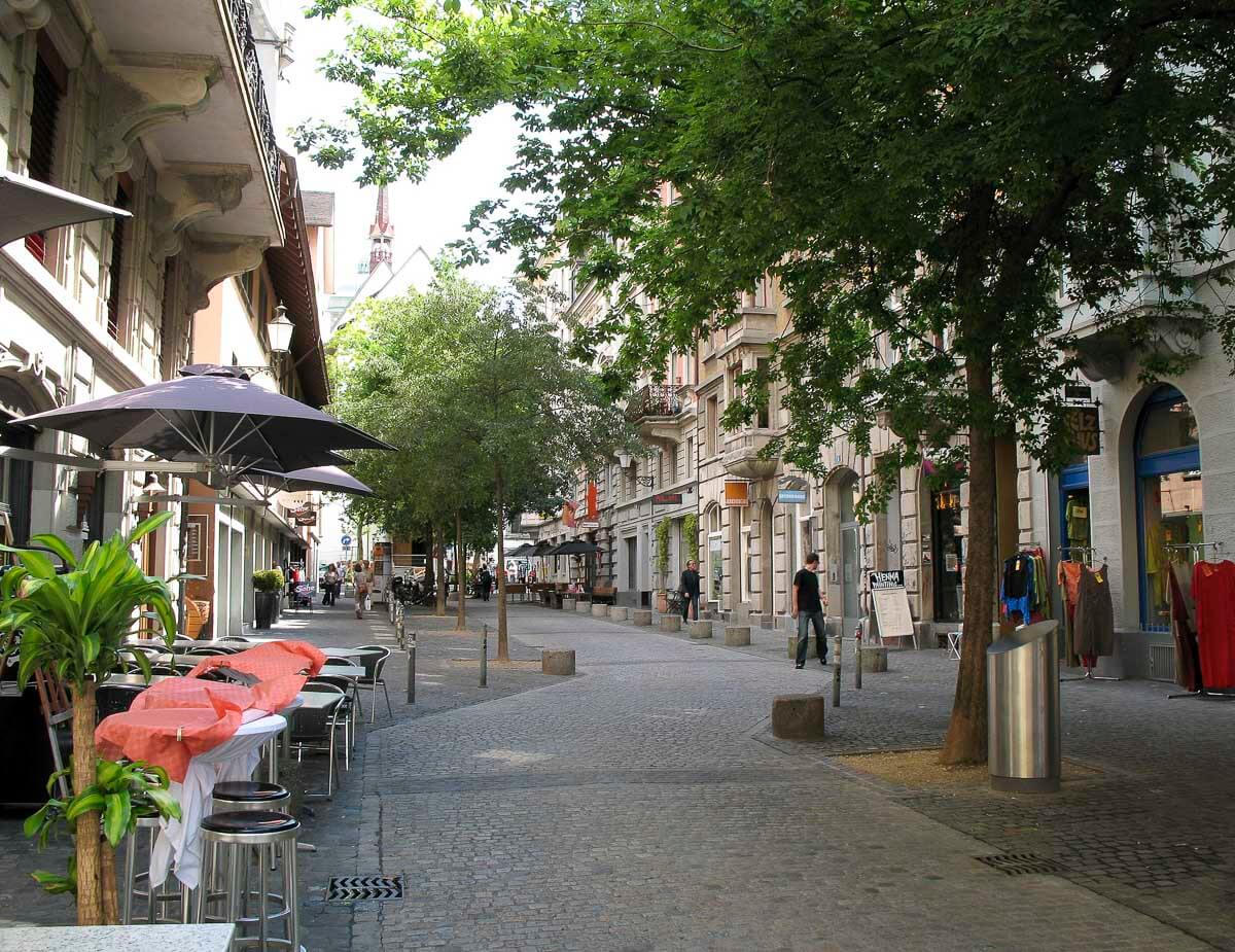 Rua charmosa e tranquila no centro de Zurique, com mesas ao ar livre e árvores sombreando o caminho.