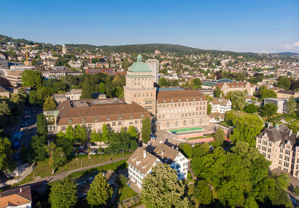 Vista aérea do prédio principal da Universidade de Zurique cercado por árvores e edifícios históricos, em um cenário urbano com colinas ao fundo.