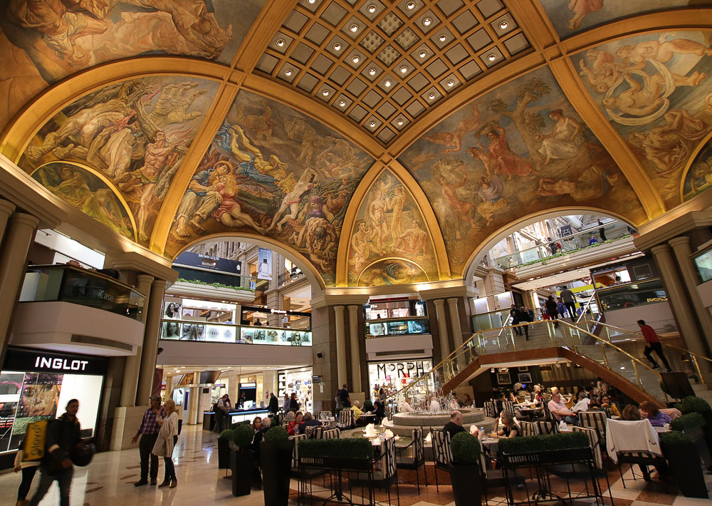 Interior das Galerías Pacífico, com teto arredondado e afrescos detalhados, além de lojas e pessoas caminhando e sentadas em um café.