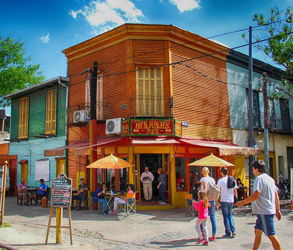 Casinhas coloridas de vermelho, verde e amarelo no bairro de La boca, com pessoas passeando em um dia de sol.
