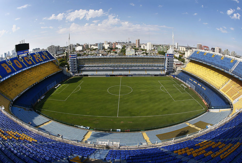 Estádio de futebol La Bombonera, em Buenos Aires, com arquibancadas azuis e amarelas e o gramado bem cuidado.