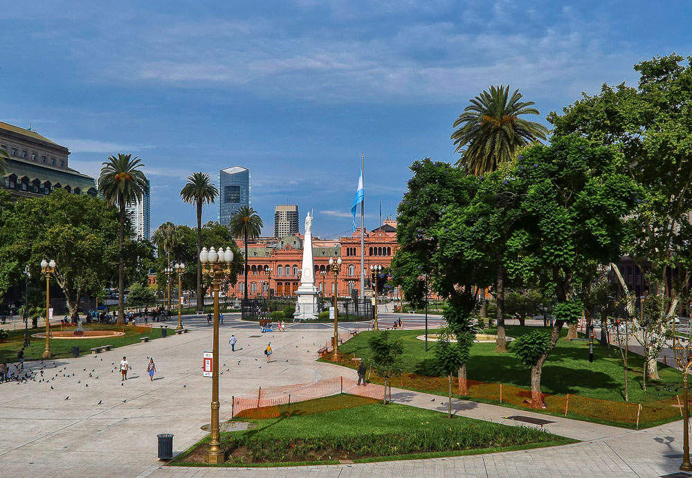 A Plaza de Mayo em um dia nublado, com palmeiras, estátuas e a Casa Rosada ao fundo.