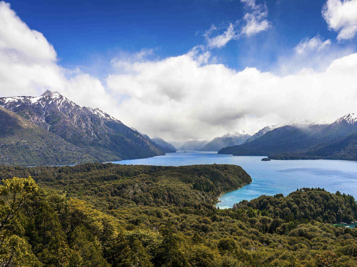 Vista panorâmica no topo do Cerro Llao Llao, com montanhas e um lago azul, cercado por florestas exuberantes.