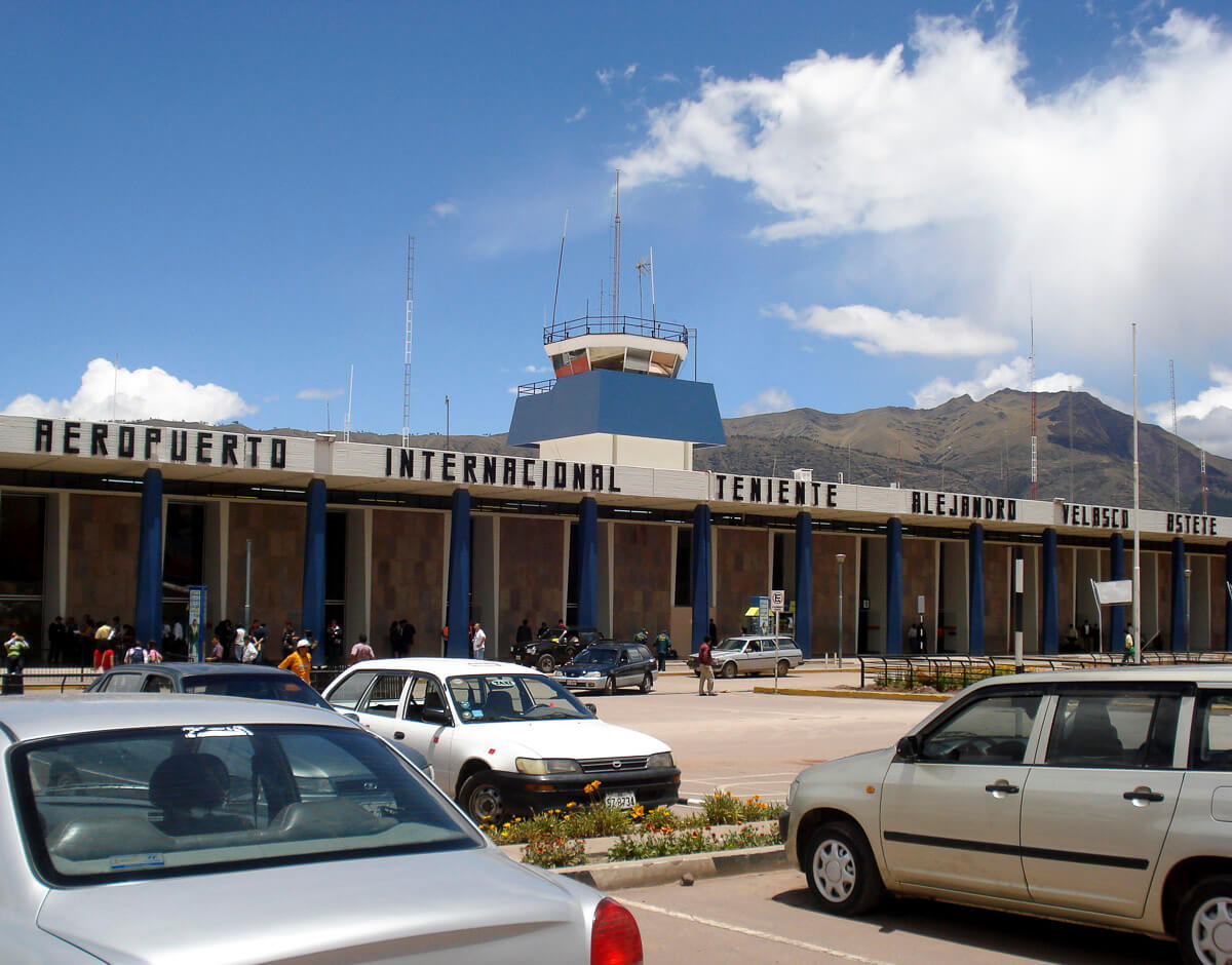 Fachada do Aeroporto Internacional Alejandro Velasco Astete, em Cusco, com montanhas ao fundo e carros estacionados na frente.