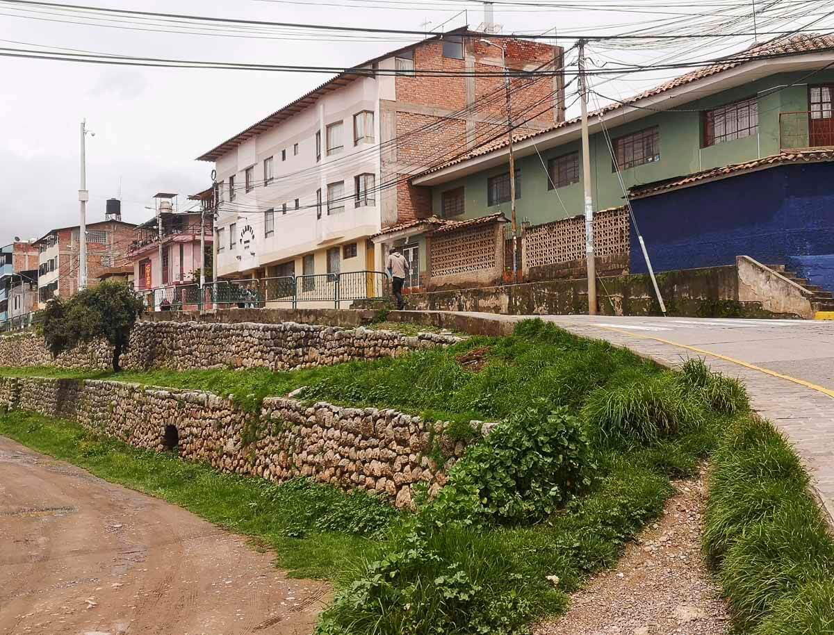 Rua residencial em Lucrepata, Cusco, com casas simples, calçadas de pedra e vegetação.