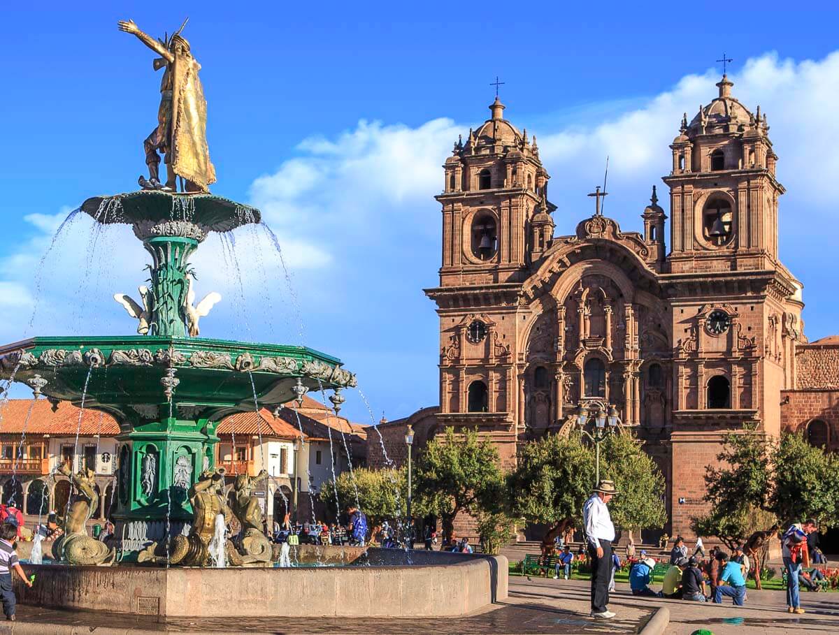 Praça principal de Cusco com uma fonte ao centro e a Igreja da Companhia de Jesus ao fundo.