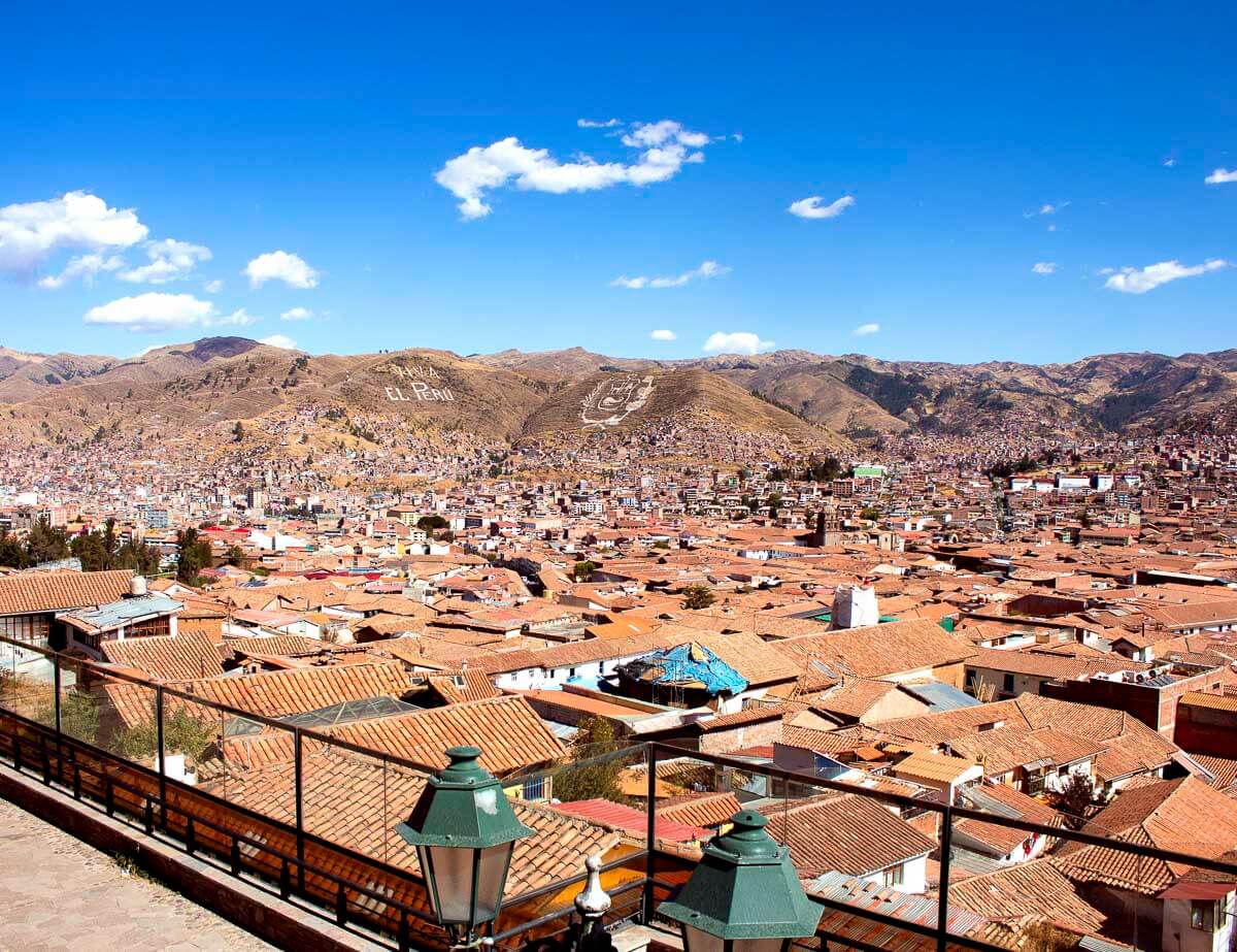 Vista panorâmica de Cusco com casas de telhado vermelho e montanhas ao fundo, destacando a palavra “El Perú” escrita em uma colina.