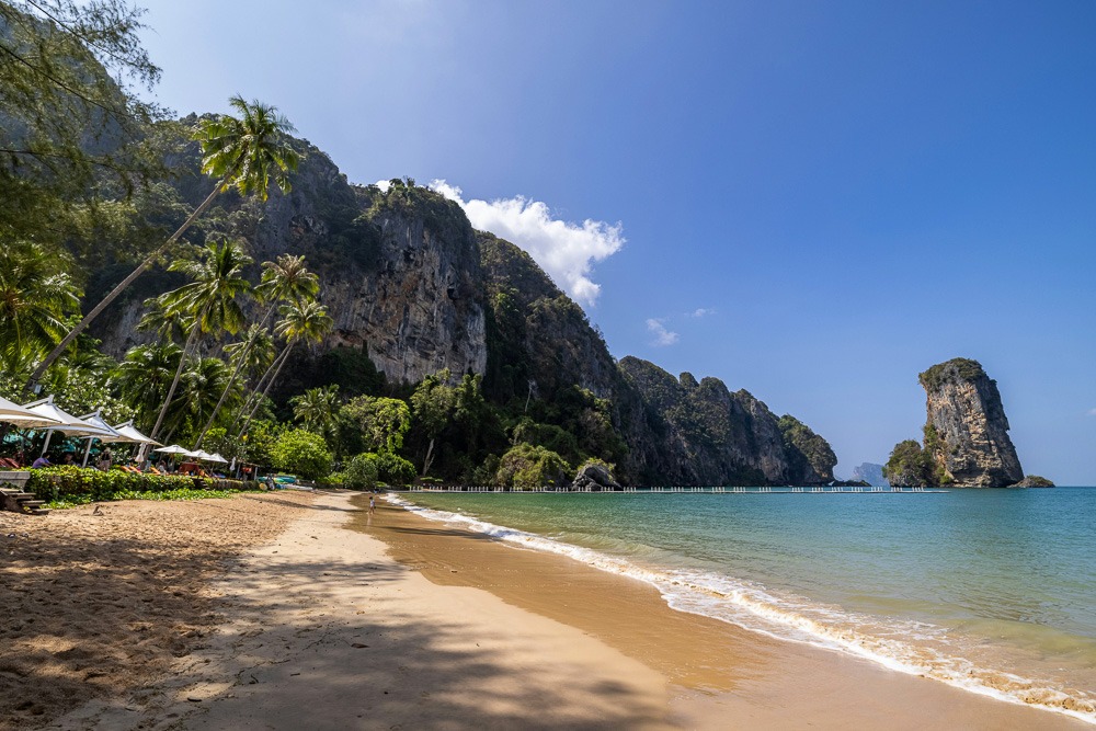 Praia de Ao Nang com areia dourada, cercada por palmeiras e montanhas calcárias ao fundo. O mar azul encontra o céu claro e ensolarado.