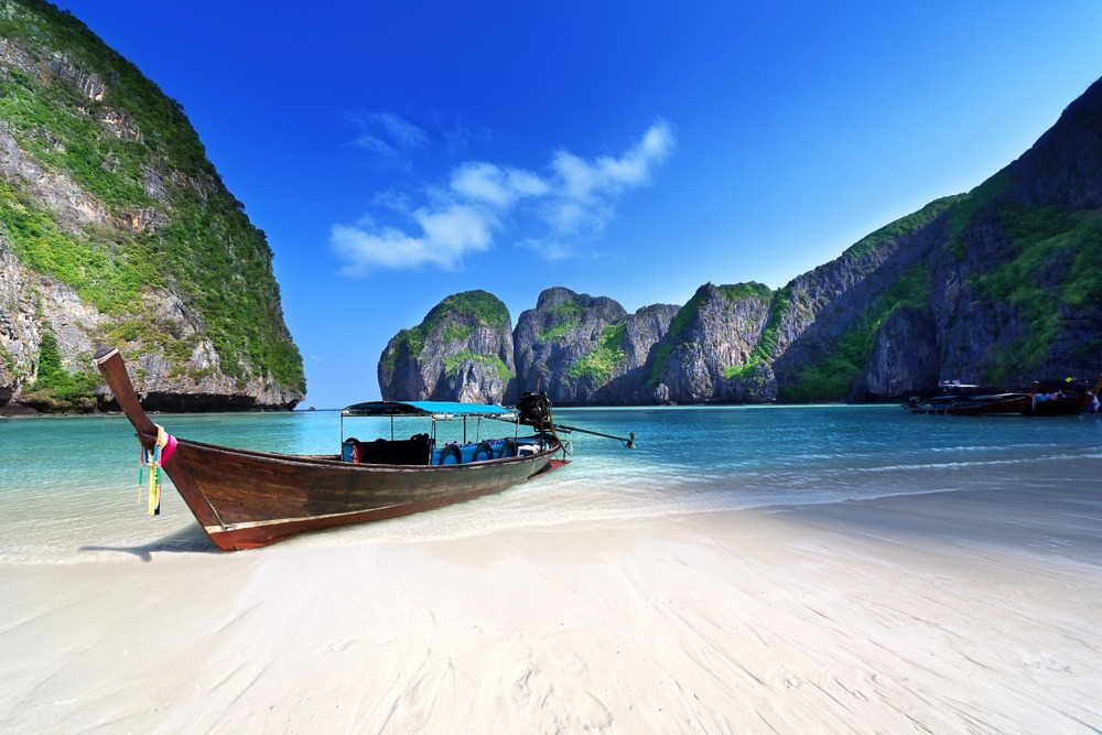 Barco de madeira tailandês ancorado em uma praia de areia branca na baía de Maya Bay, cercada por imponentes penhascos de calcário cobertos de vegetação.