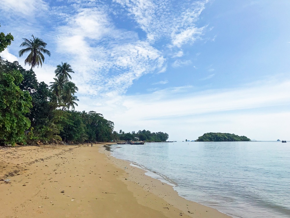 Praia de Tubkaek com vegetação tropical, areia clara e mar azul, vista de uma faixa elevada.