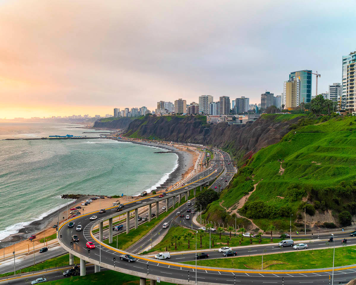 Vista aérea da orla de Miraflores, em Lima, com prédios modernos e áreas verdes ao pôr do sol.