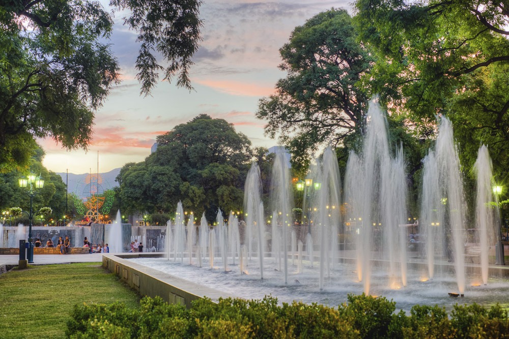 Fonte iluminada na Plaza Independencia em Mendoza, cercada por árvores e arbustos, com o céu ao entardecer ao fundo.