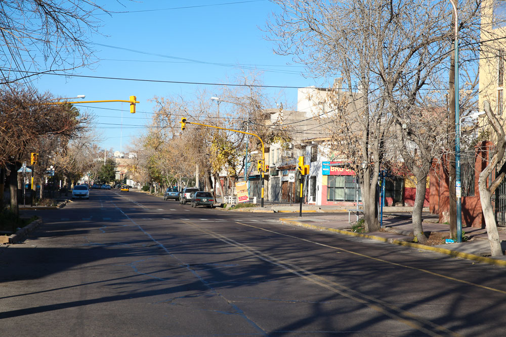 Rua tranquila em Godoy Cruz, Mendoza, com árvores sem folhas, semáforos e algumas lojas locais.