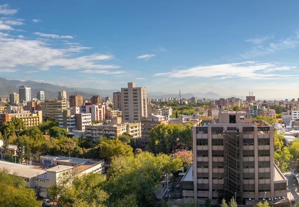 Vista aérea de Mendoza, com edifícios residenciais e comerciais cercados por árvores e montanhas ao fundo.