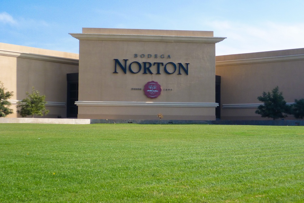 Fachada da Bodega Norton em Mendoza, com gramado verde e céu azul ao fundo.