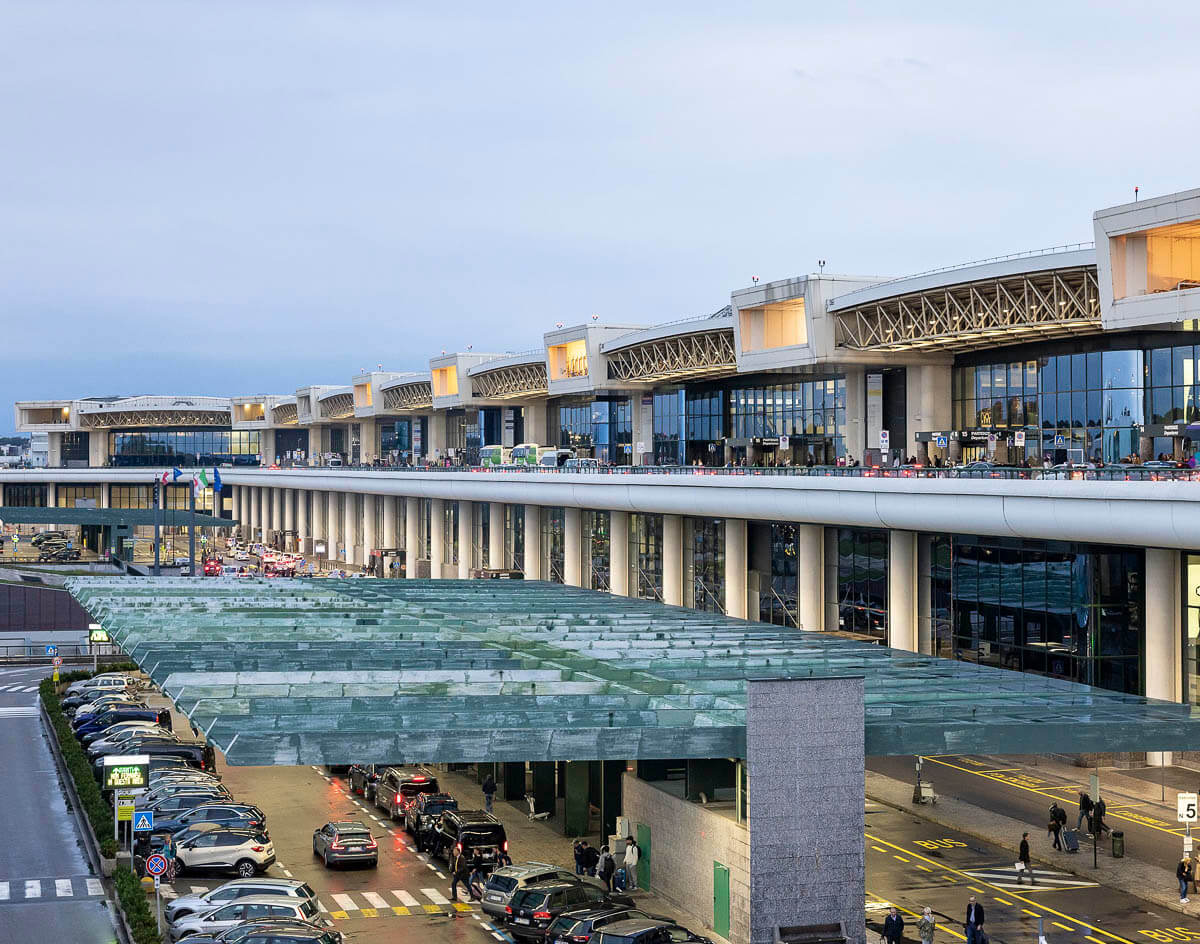 Vista do terminal principal do Aeroporto de Malpensa, em Milão, com estrutura moderna e carros no estacionamento.