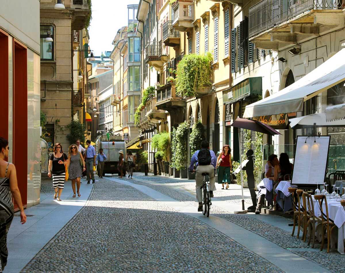 Rua charmosa do bairro Brera em Milão, com pedestres, ciclistas e mesas de restaurantes ao ar livre.