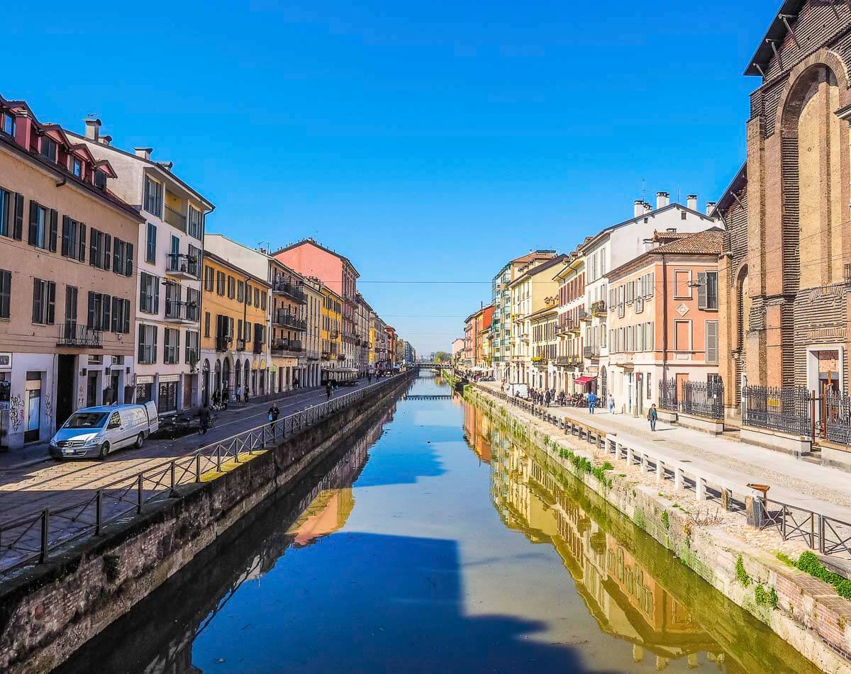 Vista do canal Naviglio Grande com prédios coloridos refletidos na água sob céu azul.