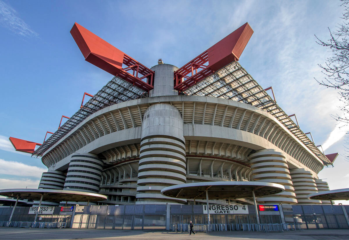 Fachada do Estádio San Siro, em Milão, com sua estrutura circular icônica e elementos em vermelho e cinza.