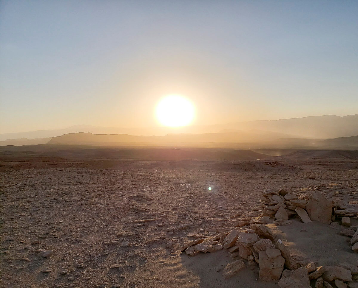 Deserto do Atacama ao pôr do sol, com luz dourada iluminando a paisagem árida e montanhas ao fundo.
