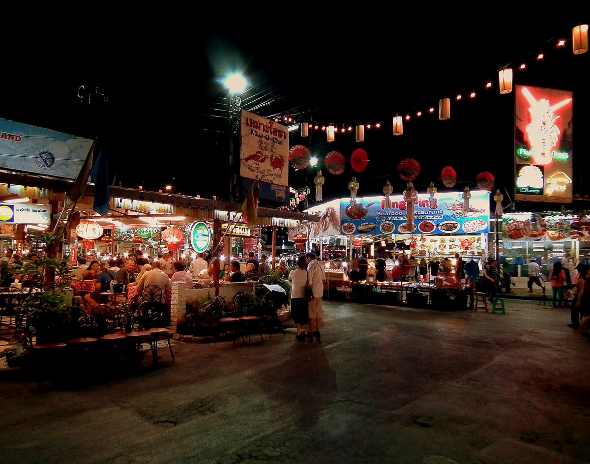 Uma movimentada feira noturna em Chiang Mai, iluminada por luzes e decorada com letreiros coloridos, com pessoas jantando em mesas ao ar livre e barracas oferecendo comida local.