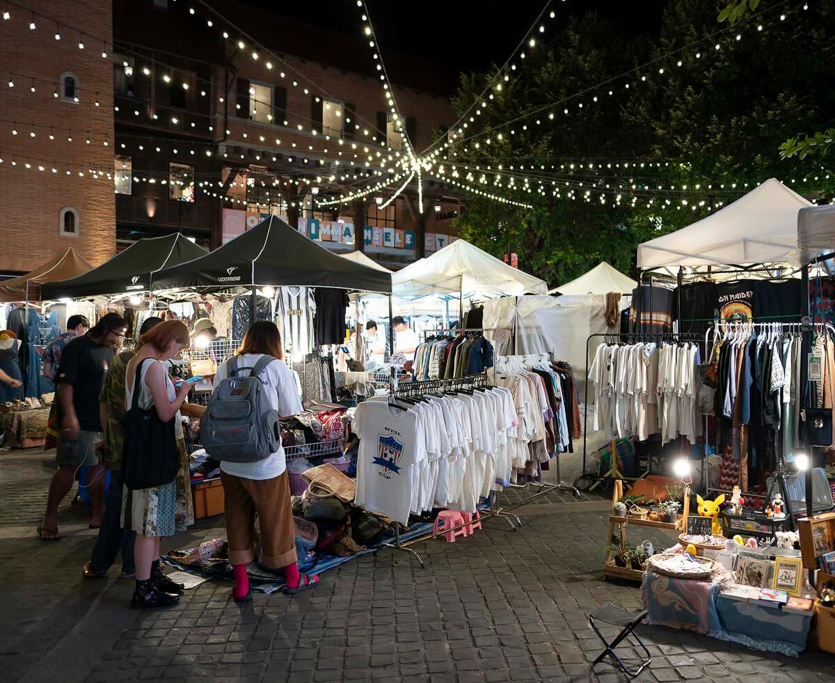 Rua movimentada em Chiang Mai com barracas de roupas e produtos artesanais, decorada com luzes penduradas e cercada por árvores e prédios.
