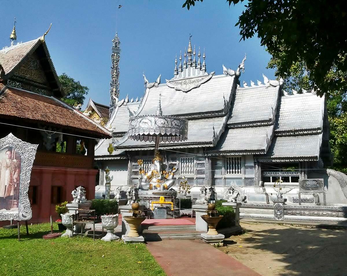 Templo budista prateado com estátuas e ornamentos detalhados, cercado por árvores e grama.
