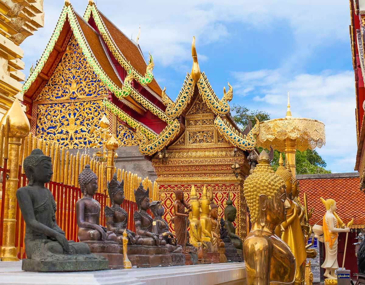Fachada do Wat Phra That Doi Suthep, um templo dourado com estátuas de Buda, cercado por ornamentos tailandeses e um céu azul ao fundo.